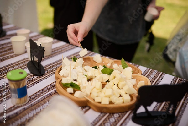Obraz People take cheese cut into cubes from a wooden plate on a tablecloth with an ornament