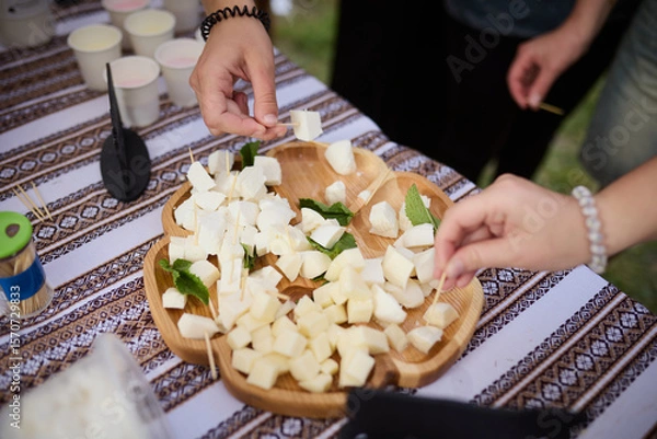 Obraz People take cheese cut into cubes from a wooden plate on a tablecloth with an ornament