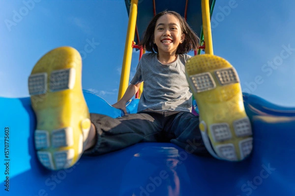 Obraz A little Asian girl playing at playground slide looking directly at the camera with a wide, joyful smile. Childhood play, fun, and excitement concept.
