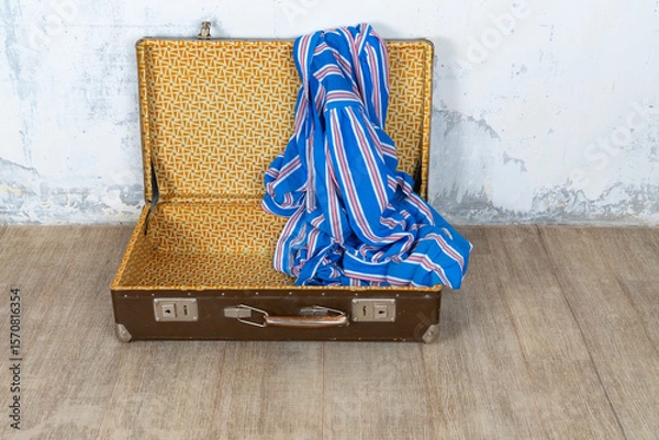 Fototapeta Vintage retro suitcase, the suitcase is open and there is a scarf in it, stand on a stool. The background is a white wall. The concept of retro travel and retro design.