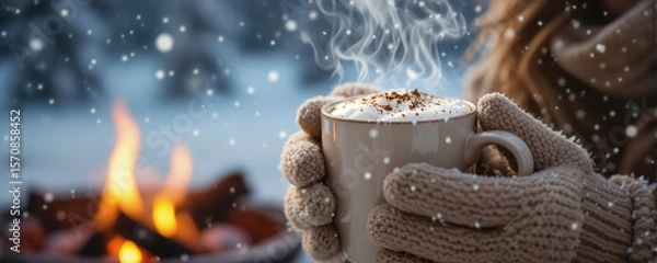 Obraz Cozy person in knitted mittens holding a steaming mug of hot cocoa by a warm campfire in a snowy winter landscape with falling snowflakes, Winter Holidays.