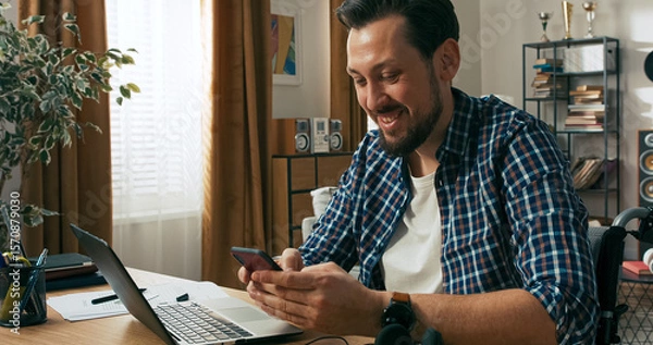 Fototapeta A bearded man with a disability multitasks from home. He writes messages on his computer and watches a video on his phone, exchanging stories with friends.