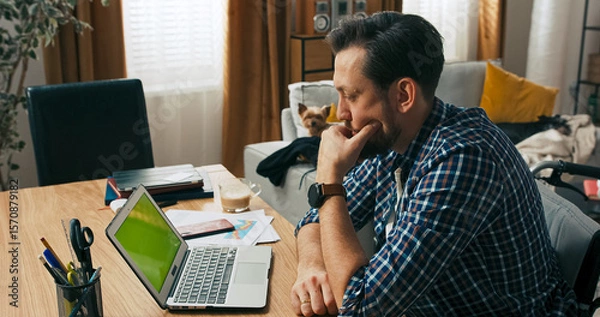 Fototapeta A concentrated man with a disability sits in a wheelchair at his desk, working online. His expression shows determination as he carefully thinks through a digital challenge.