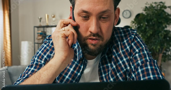 Fototapeta A serious male employee with a beard talks on the phone while working online, confused and thoughtful as he discusses challenges related to a new digital project.