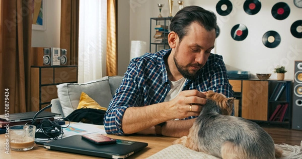 Fototapeta A happy young man in a shirt holds a credit card while shopping online, smiling with joy as a small domestic puppy sits beside him on the table, adding warmth to the moment.