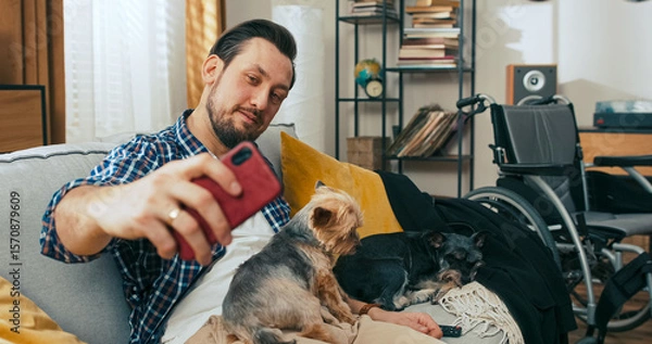 Fototapeta A man with a disability who is middle-aged relaxes on the couch, holding a phone and spending time with his dogs, enjoying comfort, connection, and calm at home.