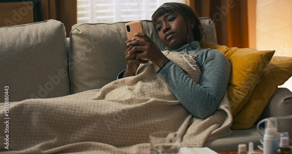 Fototapeta A woman rests quietly under a warm blanket after battling fever and a runny nose. Her bedside table holds tissues, tablets, syrups, and sprays, supporting her recovery at home.
