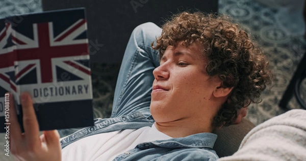 Fototapeta A teenage student prepares for a school exam, lying on the couch and reading English words aloud from a book.