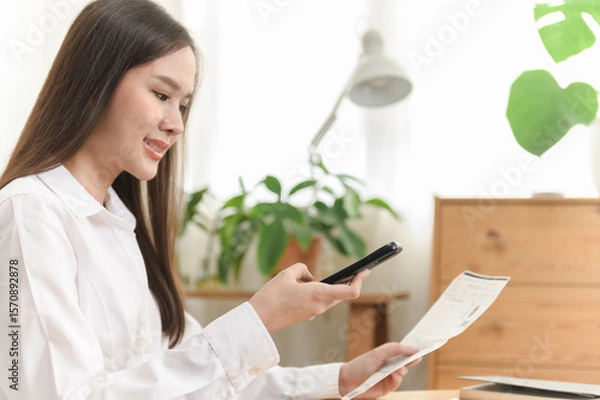 Fototapeta Young Asian woman using smartphone to scan and pay utility bill at home, demonstrating convenient electronic payment and mobile banking technology in modern lifestyle.