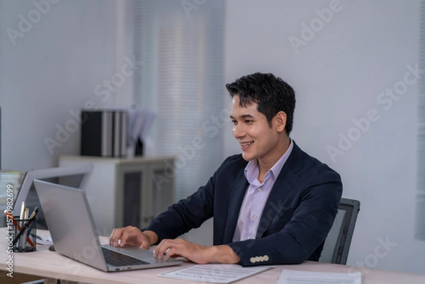 Fototapeta A man in a suit is sitting at a desk with a laptop in front of him