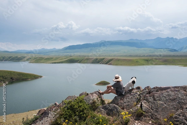 Obraz A woman and her dog friend sit on rocks above a mountain lake in summer, admiring the surrounding landscape of Kazakhstan's wild nature. Bestyubinskoye Reservoir, Bestobe on the Charyn River.