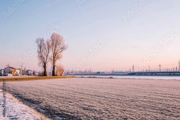 Obraz Frosty winter landscape.  Bare trees stand in a frosted field, with a river and distant city skyline in the background.  Pale sunrise colors paint the sky