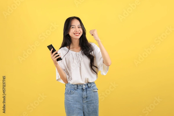 Fototapeta Young Indonesian Teenager with Long Hair Smiling with One Hand Raised While using a Black Smartphone