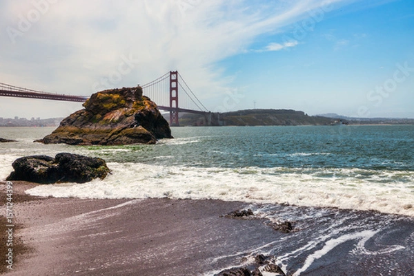 Fototapeta Golden Gate Bridge view from the rocky shoreline on a clear day in San Francisco, California. Coastal waves, iconic red architecture, and Sutro Tower in the distance capture the essence of the Bay