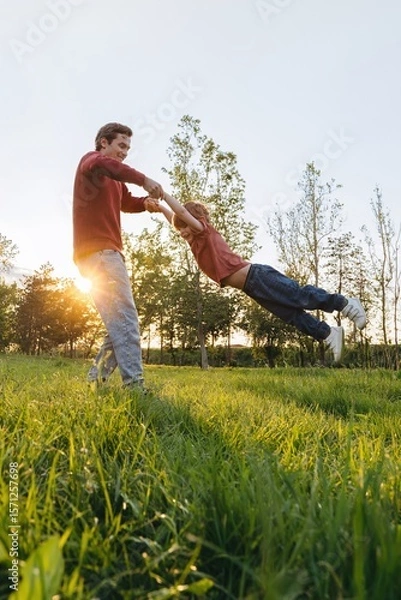 Obraz Playful young father spinning his happy son around in a grassy park during sunset. Smiling dad playing with his joyful child outdoors. Family connection and bonding. Space for text