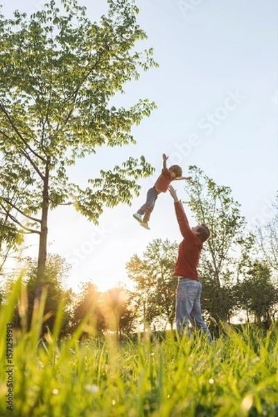 Obraz Father joyfully tossing his young son high into the air at sunset in a lush green park. Fathers day concept. Wide shot with space for text