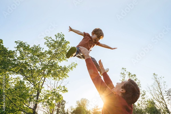 Obraz Father tossing his laughing joyful son into the air against a bright sky in a park on a sunny day. Cheerful boy playing with his dad outdoors, creating happy childhood memories together. Copy space
