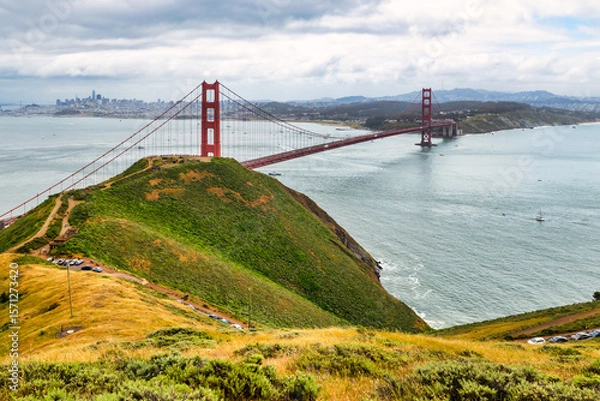 Fototapeta Panoramic view of the Golden Gate Bridge spanning San Francisco Bay with cityscape, lush hills, and boats, captured from a high vantage point on a cloudy spring day