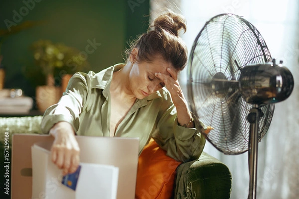Obraz A distressed woman, hand on forehead with eyes closed, battles the combined discomfort of summer heat and the overwhelming burden of financial responsibilities, indicated by bills and a fan.