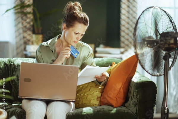 Obraz A focused woman manages home finances, reviewing documents with a credit card & laptop. Seated on a green velvet sofa, she navigates bill payments; a floor fan hints at the warm indoor environment.