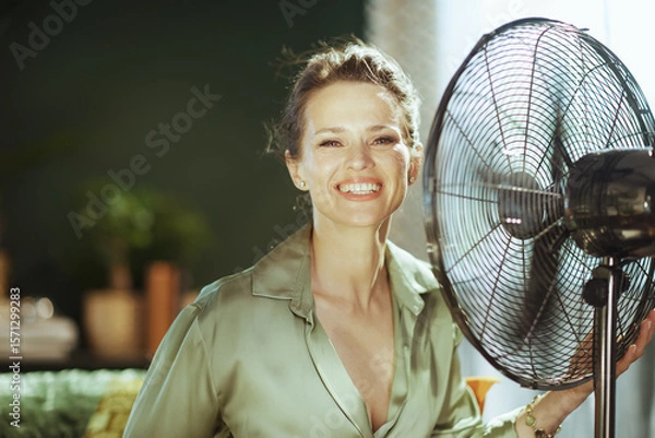 Obraz A smiling woman, refreshed by an electric fan, looks at the camera, embodying the ease of summer cooling indoors. Soft window light adds a pleasant glow to this scene of domestic well-being.