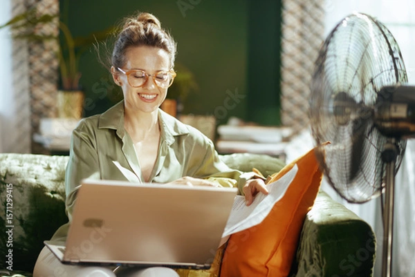 Obraz A smiling woman with glasses works comfortably from her green sofa, holding papers and using a laptop. A floor fan nearby provides relief, highlighting a productive home office setting in warm weather