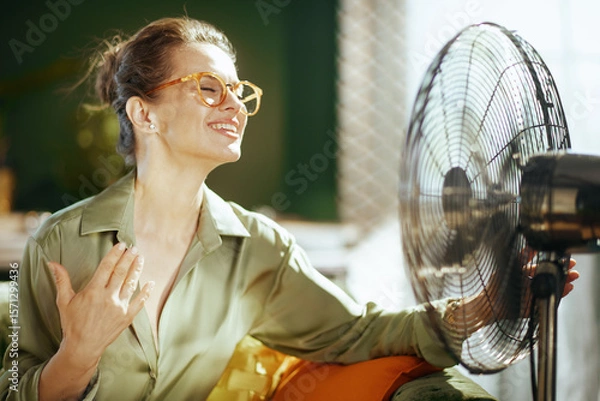 Obraz A middle-aged woman, eyes closed & smiling, tilts her head back, enjoying the refreshing breeze from a fan. Her hand on her chest conveys relief & comfort from summer heat in her indoor setting.
