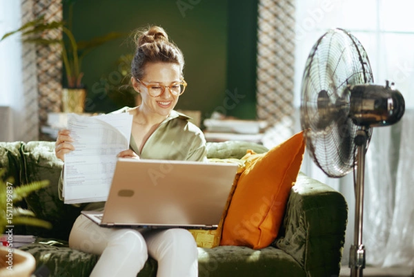 Obraz A smiling woman with stylish glasses and a messy bun sits comfortably on a green sofa, working on her laptop. A fan in the background suggests a warm, productive summer day in her home office.