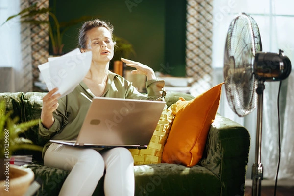 Obraz A middle-aged woman on a green velvet couch, fanning herself with papers, shows discomfort. A fan nearby emphasizes her struggle with hot indoor conditions while working.