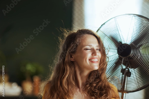 Obraz A woman with windswept brown hair smiles with closed eyes, standing before a metal fan. This indoor scene conveys comfort, relief, and enjoyment in warm weather.