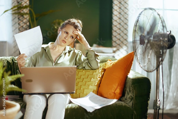 Obraz A distressed woman in her home office, fanning herself with papers and an exasperated hand on her forehead, struggles with heat and overwhelming work, beside a metallic fan.