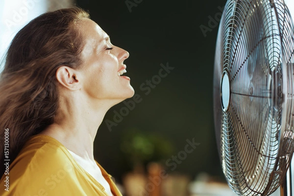 Obraz A serene woman with closed eyes and hair gently blown by an electric fan finds ultimate relief from the heat. This image captures pure comfort and refreshment during hot weather indoors.