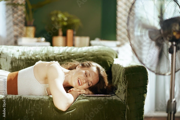 Obraz A smiling woman with closed eyes relaxes on a green velvet sofa, enjoying an electric fans cool breeze. The sunlit living room, with plants, creates a serene atmosphere of comfort and self-care.