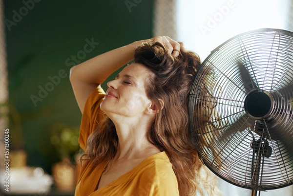 Obraz A peaceful middle-aged woman smiles with closed eyes, head tilted back, as she enjoys the refreshing breeze from an electric fan. This scene conveys comfort, relief, and well-being from summer heat.