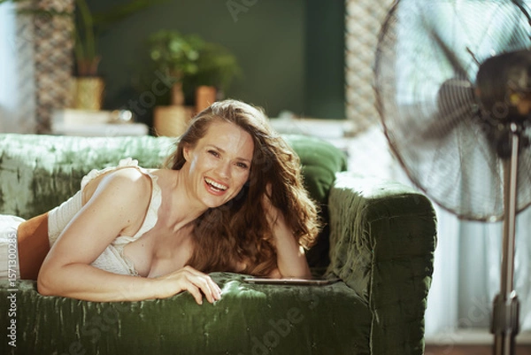 Obraz An adult woman on a green velvet sofa laughs joyfully at the camera, embodying comfort & relaxation. With a tablet & a fan cooling the bright indoor space, its a scene of pure contentment.
