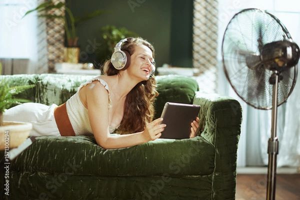 Obraz A young adult woman, smiling brightly, lies on a green velvet couch with over-ear headphones, engrossed in her tablet. A standing fan completes this relaxed scene of joyful home entertainment.