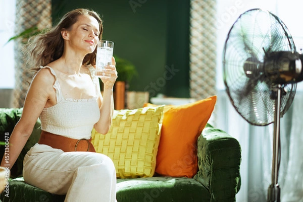 Obraz A content woman with eyes closed relaxes on a green velvet sofa, enjoying a glass of iced water and the breeze from a floor fan, finding comfort and relief from the summer heat indoors.