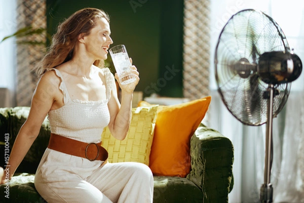 Obraz A cheerful woman with a wide smile relaxes comfortably on a green velvet couch, enjoying a cool glass of iced water and the breeze from a standing fan, finding refreshment from warm weather.