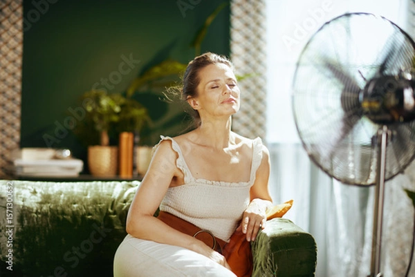 Obraz A serene woman finds comfort and relief on a green velvet sofa, eyes closed as she basks in the cooling breeze from a standing fan in her brightly lit, modern living room.