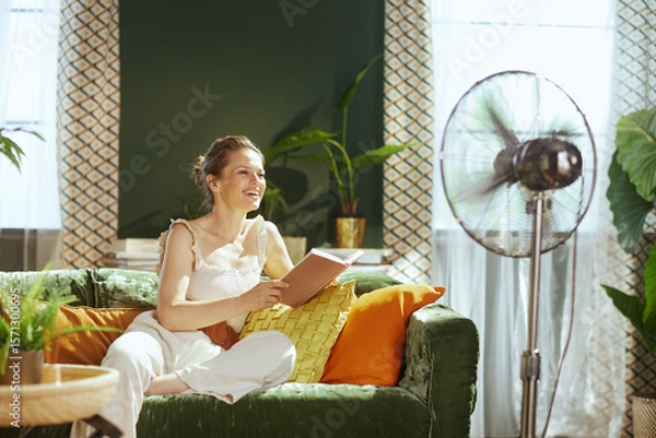 Obraz A smiling woman in a stylish living room laughs while enjoying a book and the refreshing breeze from a vintage standing fan. Bright natural light illuminates her moment of leisure and heat relief.