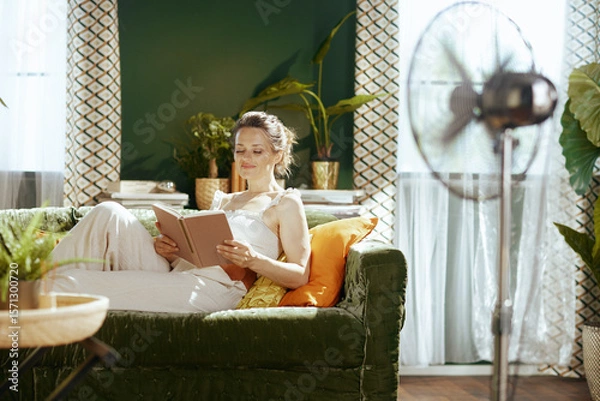 Obraz A woman with a gentle smile reclines on a green velvet sofa, eyes closed, holding an open book, lost in deep relaxation. A standing fan ensures comfort for her peaceful summer afternoon indoors.