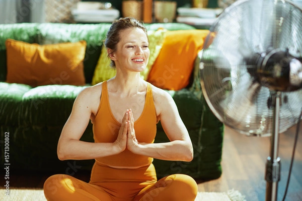 Obraz A smiling woman in orange activewear sits cross-legged, hands in prayer, enjoying a serene yoga or meditation in her living room. A spinning fan provides a cooling breeze for comfort on a warm day.