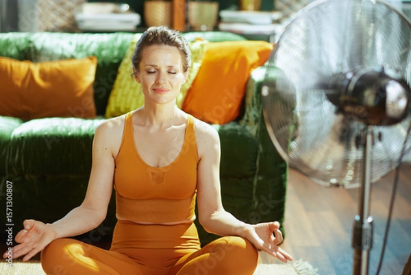 Obraz A woman in orange activewear finds tranquil serenity in a meditation pose, eyes closed and hands in mudra, benefiting from the cooling breeze of a standing fan in her sunlit living room.