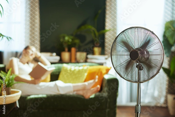 Obraz A young adult woman enjoys a book on a dark green velvet sofa in a cozy living room, complemented by a silver pedestal fan providing a cool breeze, creating an atmosphere of leisure and indoor comfort