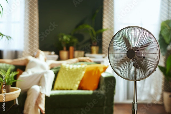 Obraz A metallic standing fan with rotating blades is in sharp focus, while a blurred woman relaxes on a green velvet sofa in a sunlit living room, evoking a sense of comfort and relief from summer heat.