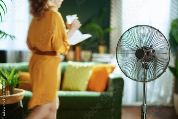Obraz A blurred woman, seen from behind, in a yellow dress holds a cup & papers in a bright living room, while a sharp metallic stand fan in the foreground provides cooling, portraying a calm domestic scene