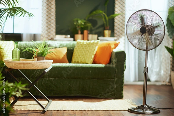 Obraz A bright & stylish living room interior with a dark green velvet sofa, vibrant yellow & orange pillows, a decorative side table, & a modern metallic standing fan, creating a refreshing atmosphere.