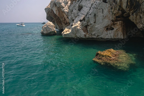Fototapeta Yacht holidays in the Mediterranean Sea. In the sea, near the rocks, people relax on yachts. A sea boat is visible next to the rock. In summer, people relax on a yacht in the sea next to large rocks.