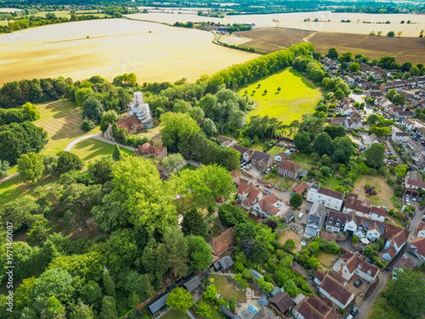 Fototapeta Pretty aerial view of a typical rural British village seen within lush trees and prime arable land. A church tower in the distance can be seen being renovated.