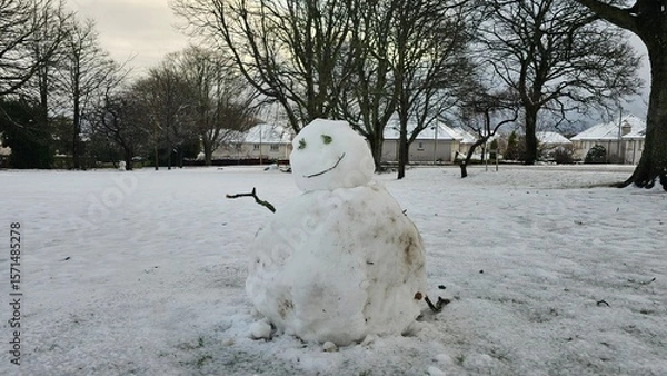 Fototapeta Smiley melting snowman on the snow with two wood stick with trees in the background